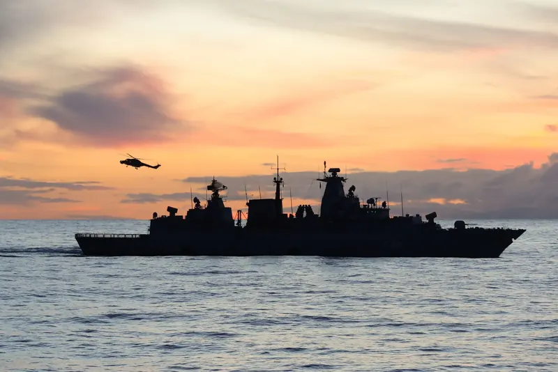 A warship and a helicopter silhouetted at sea at sunset.