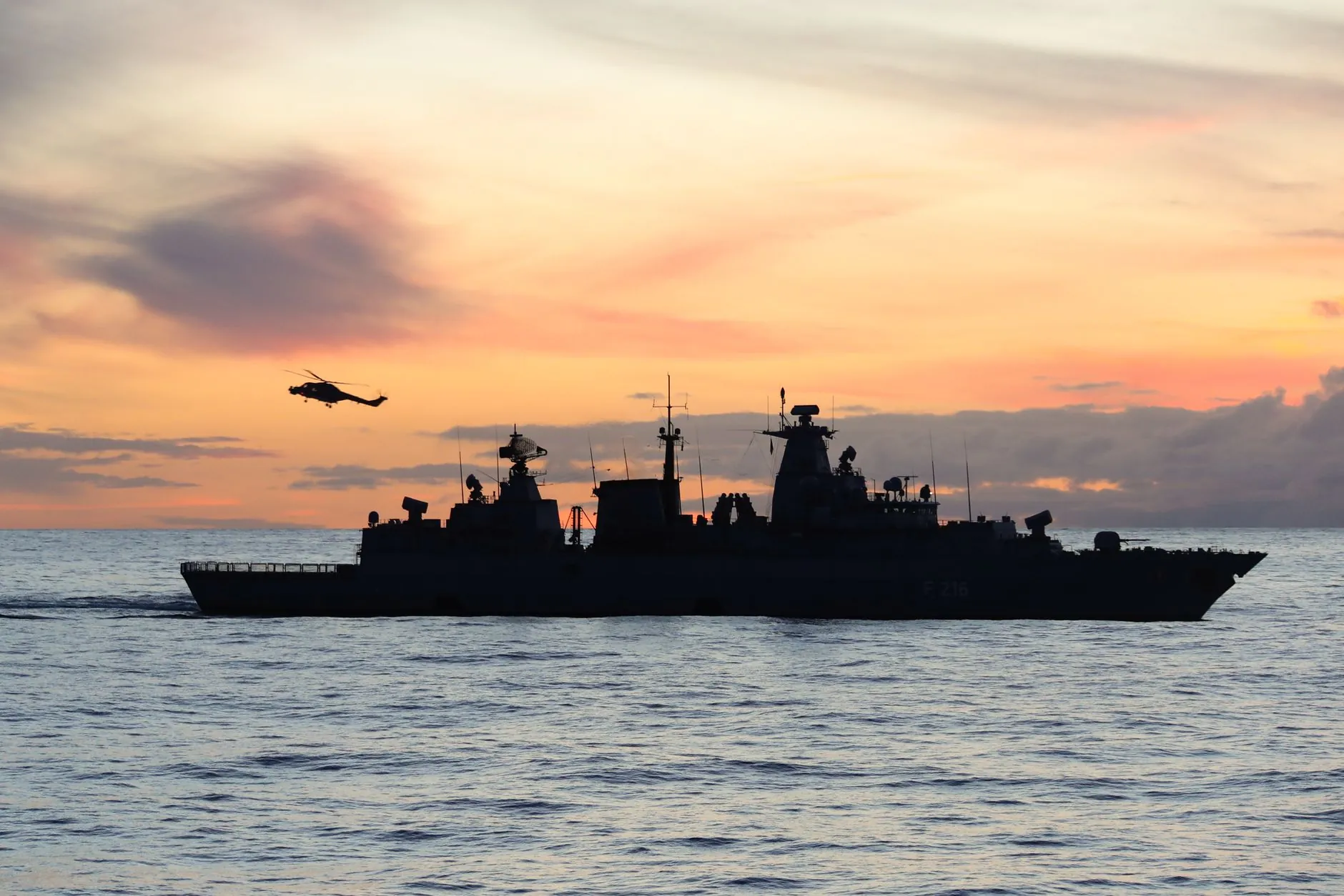 A warship and a helicopter silhouetted at sea at sunset.